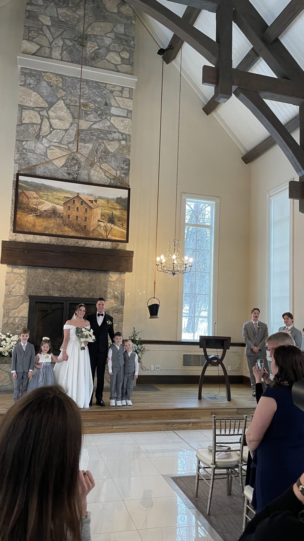 Sara and her groom with their flower girl and ring bearers at the altar of the Ancaster Mill chapel, under a tall stone fireplace and timber beams