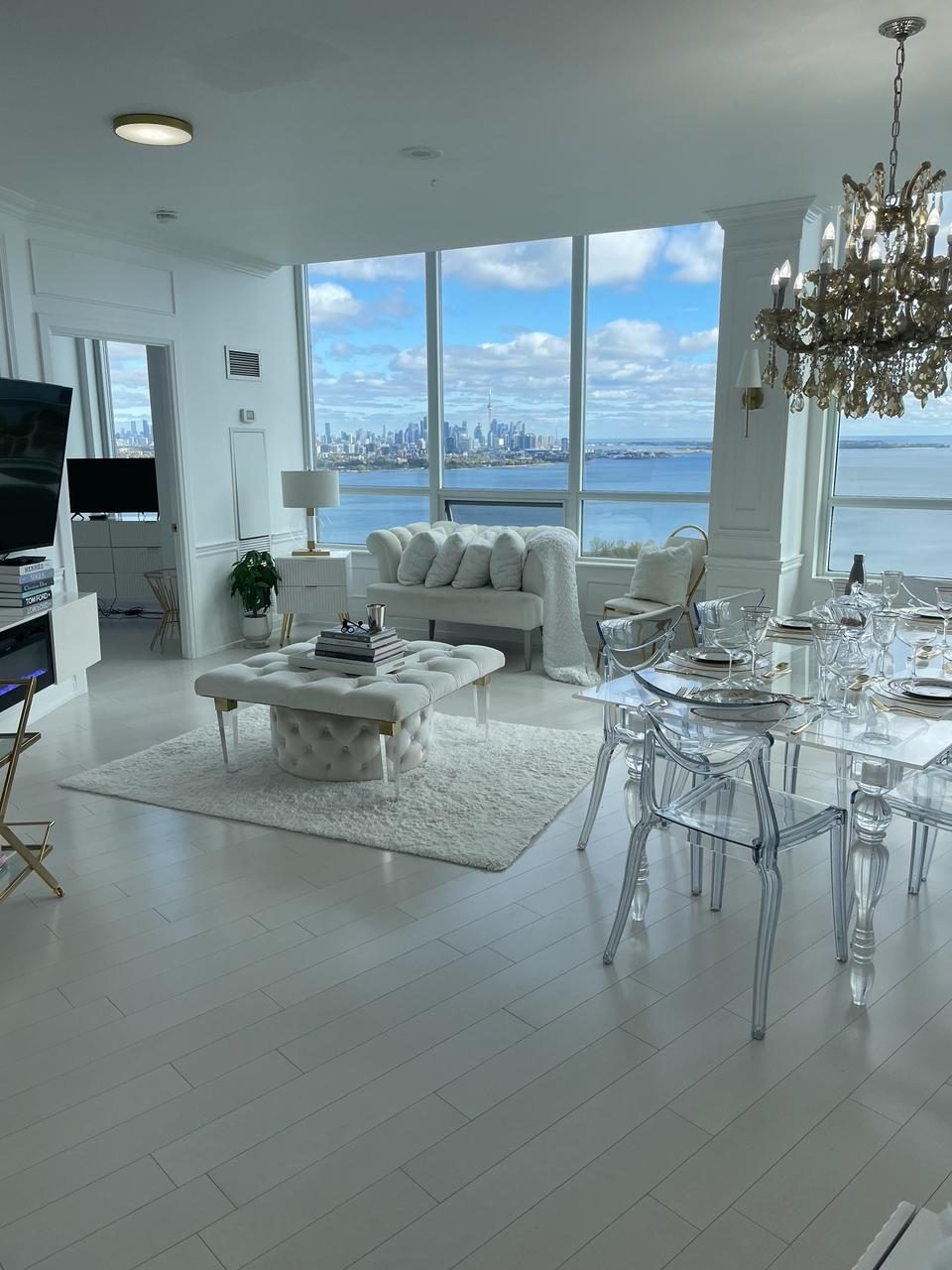 Bright white living and dining room with a tufted ottoman, a scalloped loveseat, a clear dining table with a crystal chandelier, and floor-to-ceiling windows over the Toronto skyline and the lake