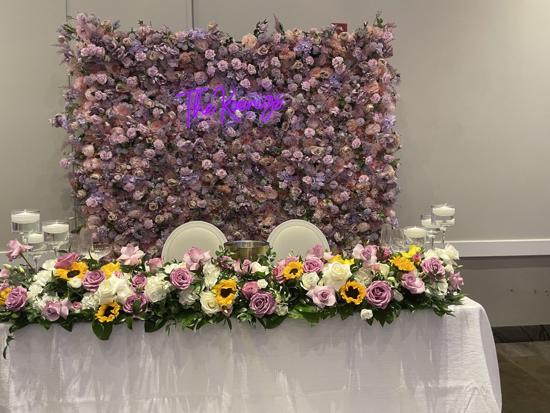 Sweetheart table with a lilac and pink flower wall, a garland of roses and sunflowers, and floating candles