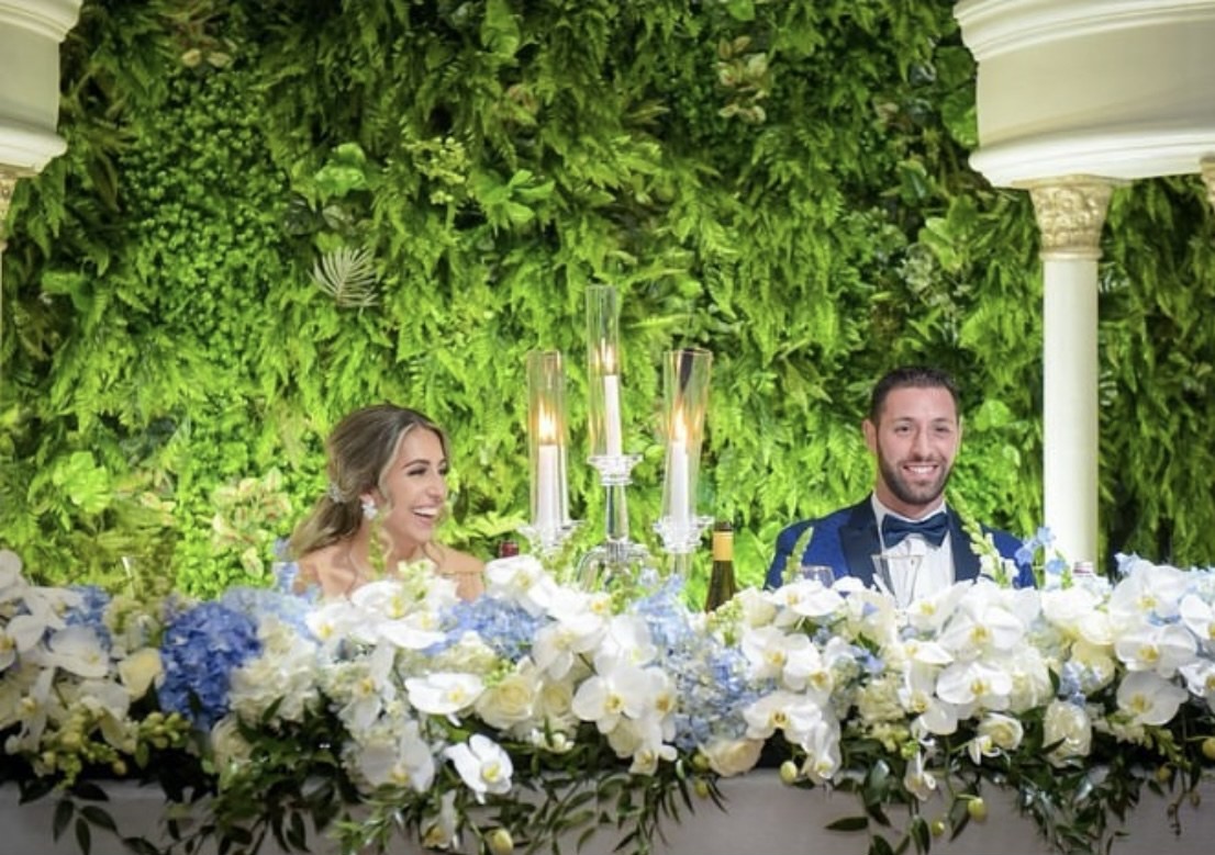 Jessica and Massimo at their sweetheart table dressed with white orchids, blue hydrangea and candelabra in front of a living green wall